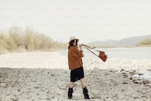 happy stylish hipster traveler woman holding bag, in hat and fringe poncho back view, walking near river, gypsy boho girl. wanderlust summer travel. atmospheric moment. space for text.
