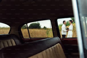 unusual view of stylish bride and happy groom through red retro car on the background of nature
