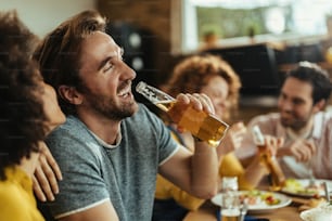 Young happy man having fun while drinking beer and eating with friends at dining table.