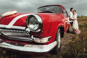 Novia con estilo y novio feliz cerca del coche retro rojo en el fondo de la naturaleza