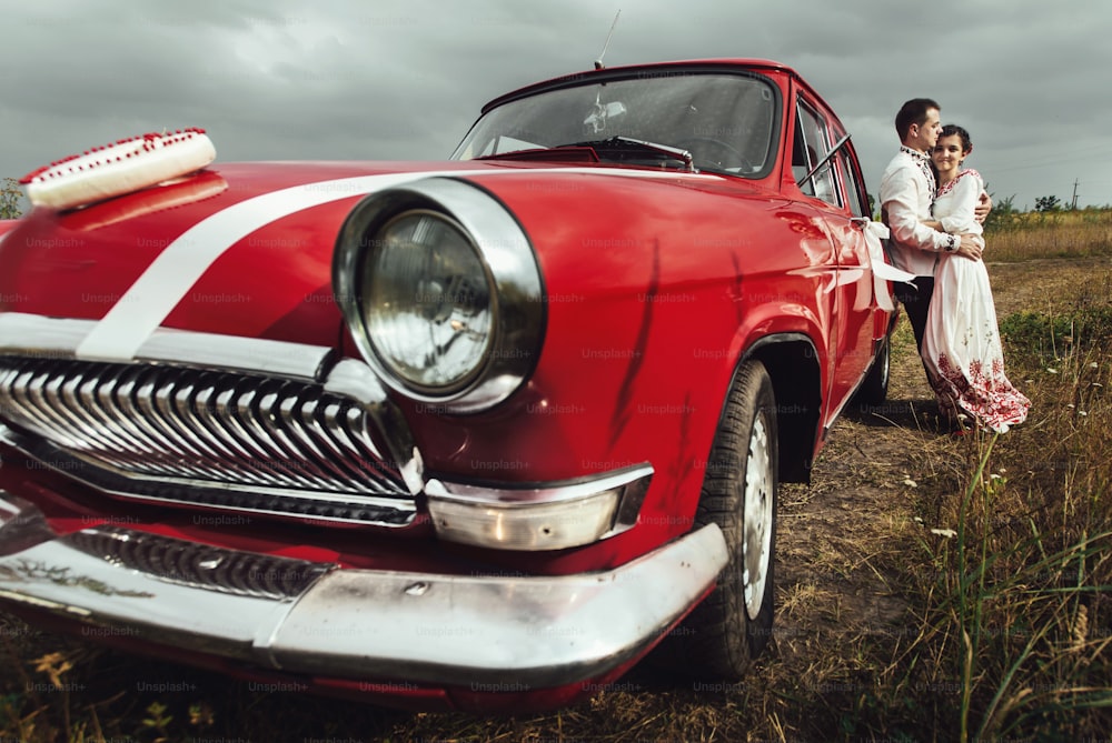 stylish bride and happy groom near red retro car on the background of nature