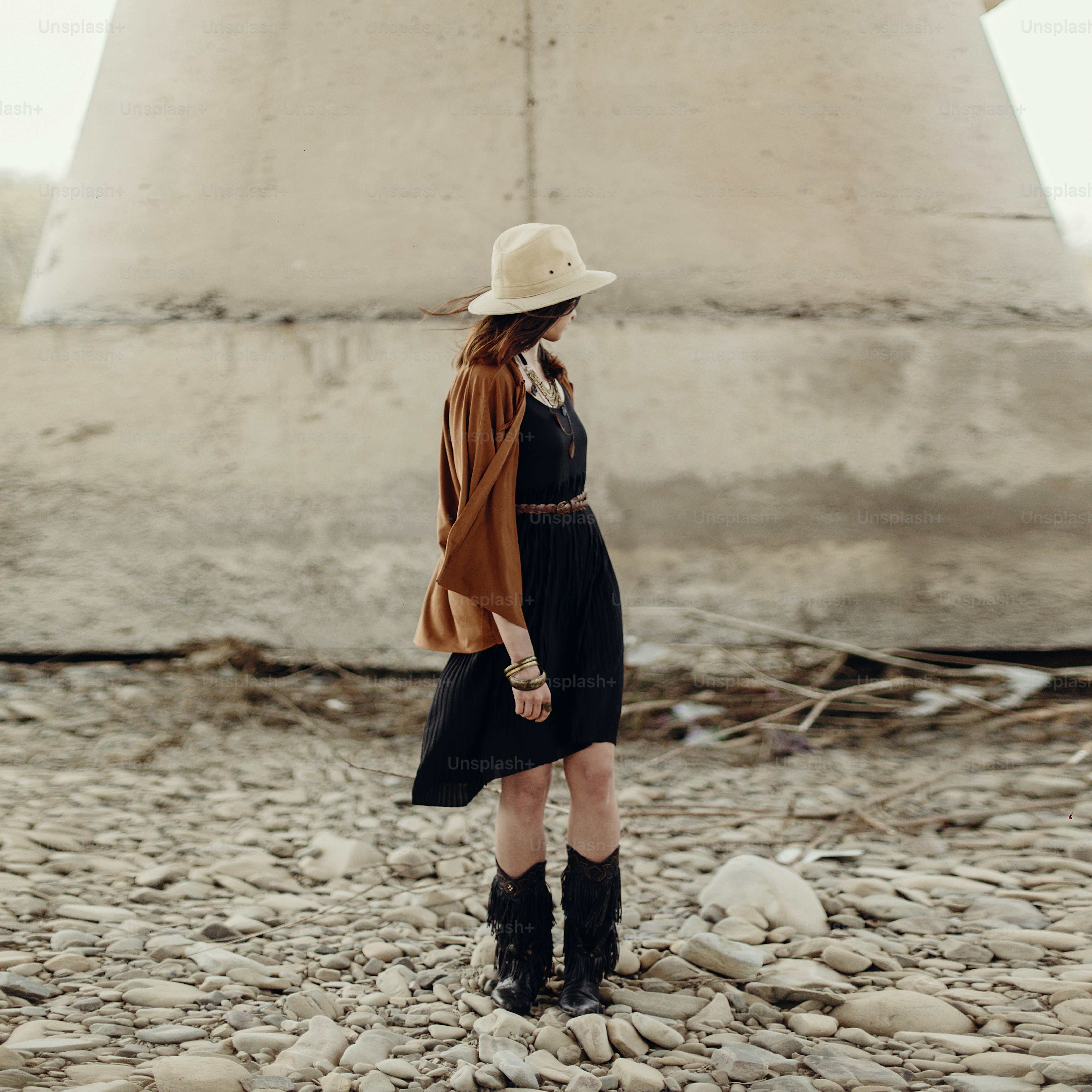 帽子、フリンジポンチョ、ブーツを持つ美しいスタイリッシュな自由奔放に生きる女性。ジプシーヒッピーの女の子は、山の川の岩の近くでポーズをとっている若い旅行者に見えます。官能的な外観。雰囲気のある瞬間。テキスト用のスペース