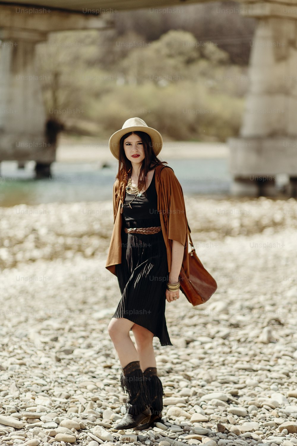 stylish boho gypsy woman in hat, fringe poncho and boots walking on river beach. hipster traveler girl look, summer travel. atmospheric moment. space for text.