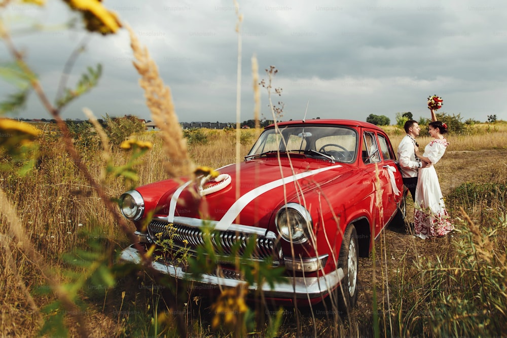 自然の背景に赤いレトロな車の近くにスタイリッシュな花嫁と幸せな花婿