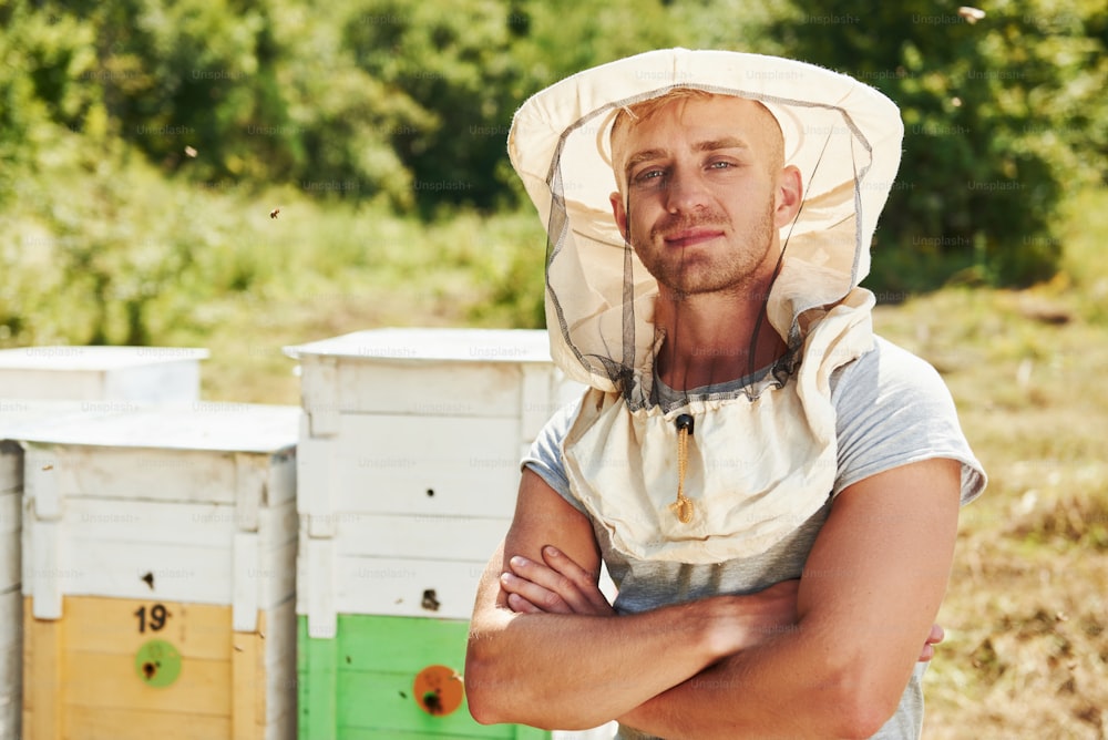 Stands with arms crossed. Beekeeper works with honeycomb full of bees outdoors at sunny day.