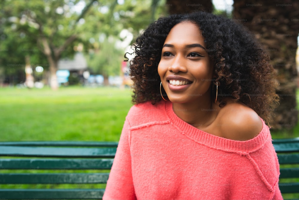 Retrato da jovem mulher afro-americana bonita sentada no banco no parque. Ao ar livre.