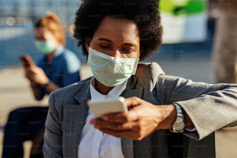 Donna d'affari nera con maschera protettiva sul viso che usa il telefono cellulare e i messaggi di testo per strada.