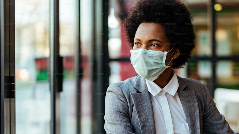 African American businesswoman wearing protective mask on her face while sitting in a cafe and looking through the window.