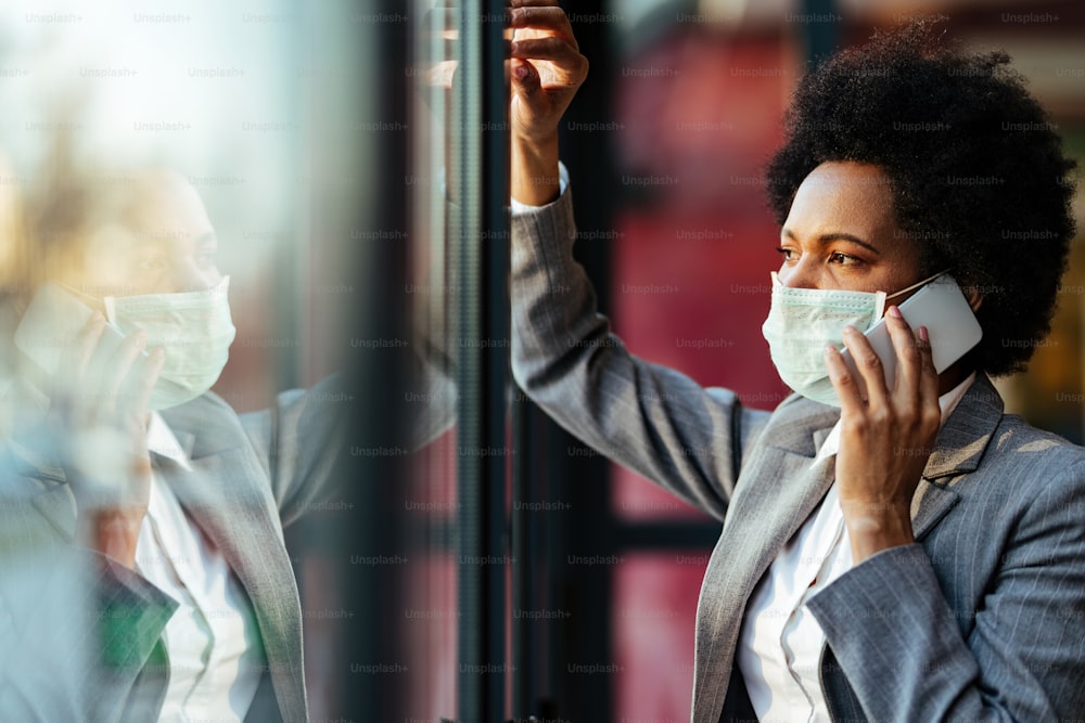 Empresária afro-americana pensativa se comunicando no celular enquanto usa máscara facial de proteção e olha pela janela.