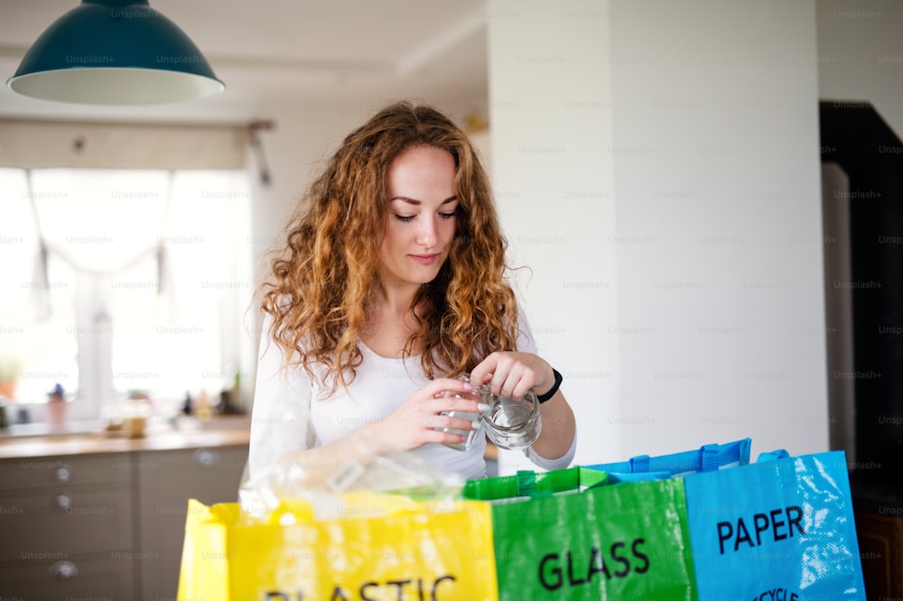 Front view of young woman indoors at home separating glass, paper, and plastic waste.