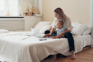 Young mother with her child working on laptop in bedroom at home. Multi-tasking, freelance and motherhood concept