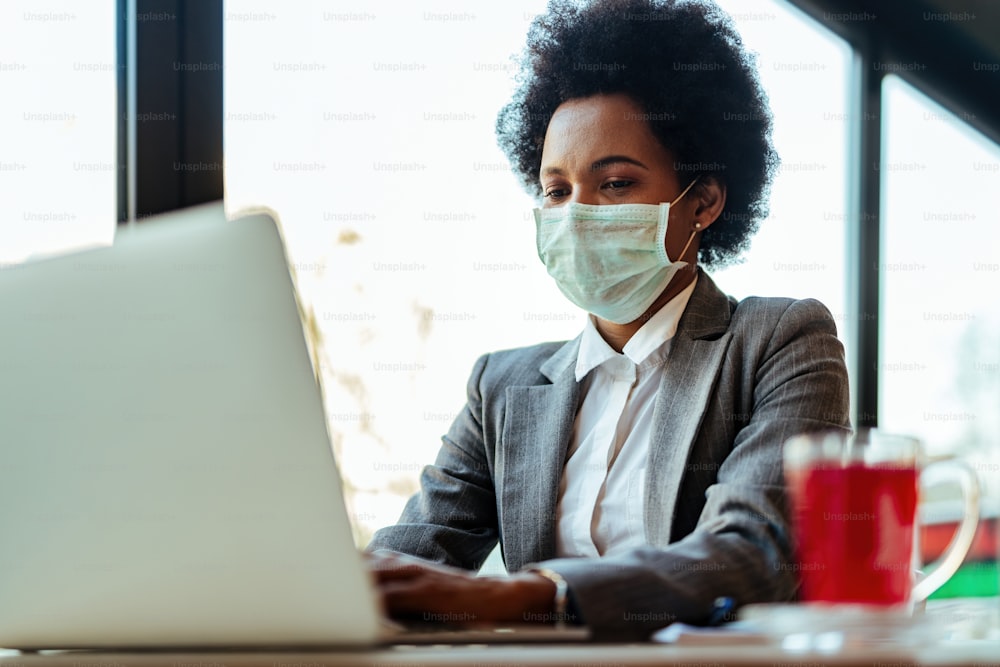 Empresaria afroamericana con máscara protectora en la cara trabajando en una computadora portátil mientras está sentada en un café.