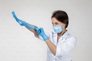 Junge Ärztin in weißer Uniform mit Maske und Gummihandschuhen auf grauem Hintergrund