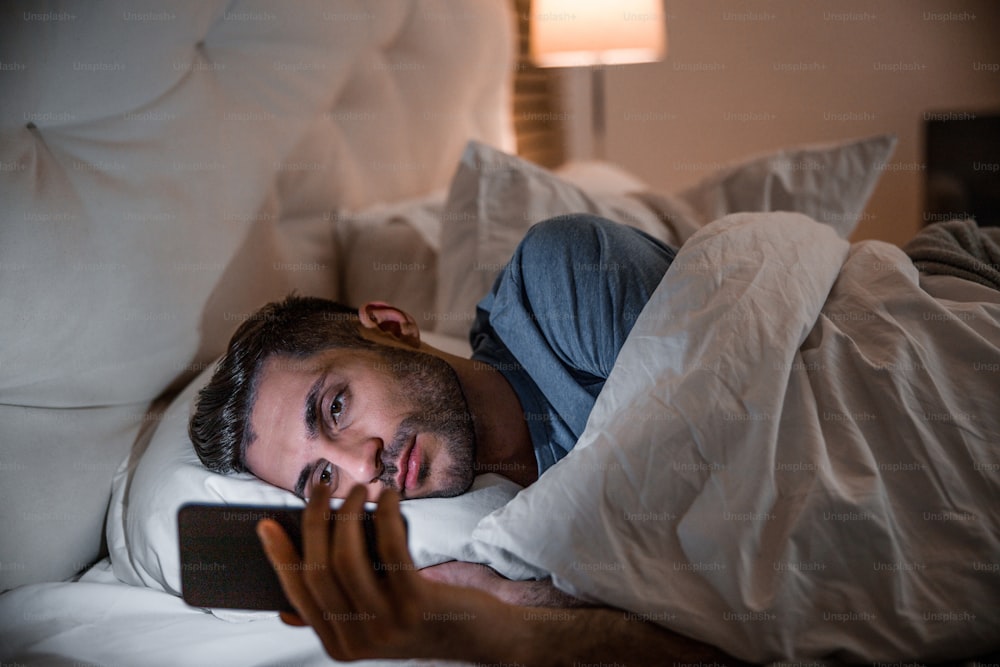 Young man is lying in comfortable bed and texting on smart phone before falling asleep
