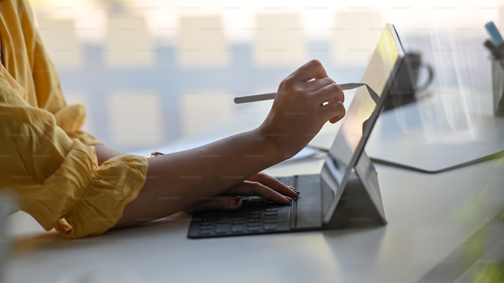 Abgeschnittenes Bild der Hand der Designer-Frau mit einem Stift zum Zeichnen auf einem Computer-Tablet mit Tastaturgehäuse, das auf einem weißen Arbeitstisch über einem komfortablen Büro als Hintergrund steht.
