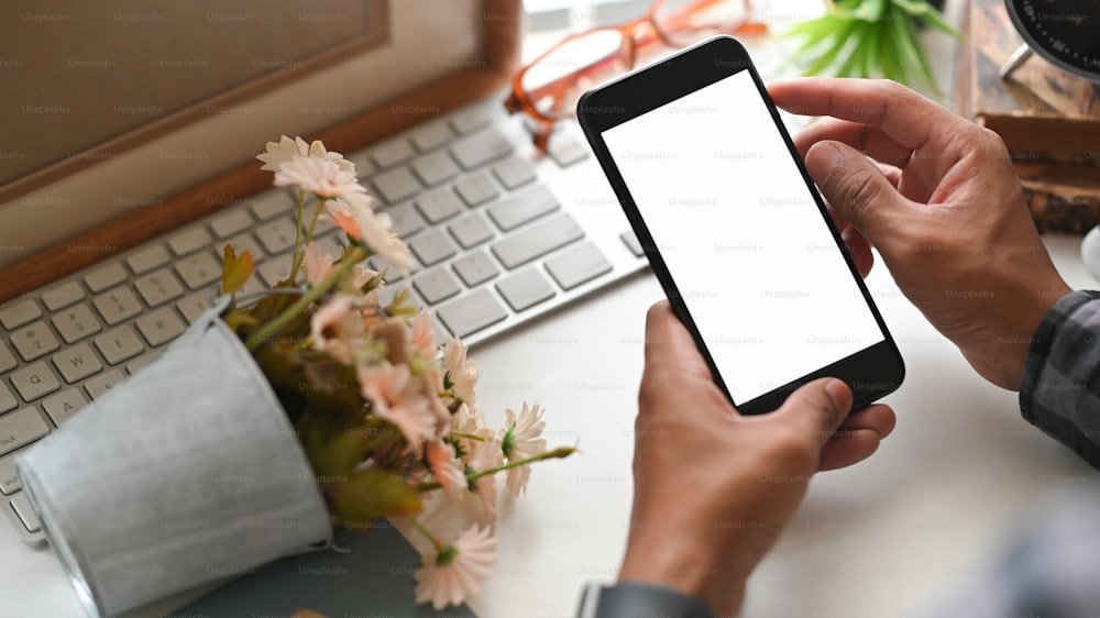 Cropped image of creative man's hands holding a cropped black smartphone with white bank screen over the wireless keyboard and bunch of flowers that putting on the white working desk.