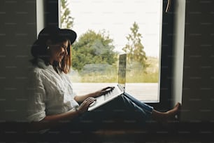 Young happy woman in hat using laptop, shopping or working online from home. Stylish hipster girl with laptop sitting on floor at big window with view into woods. Freelance and freelancer