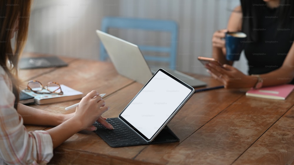 Cropped image of creative woman holding a stylus pen while drawing/pointing on white blank screen computer tablet with keyboard case that putting on working desk over comfortable room as background.