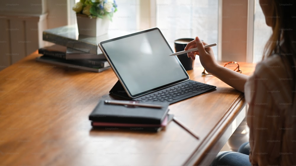 背景として快適な部屋の上の作業机に置くキーボードケース付きの白い空白の画面のコンピュータータブレットに描画/指しながら、スタイラスペンを保持している創造的な女性のトリミングされた画像。