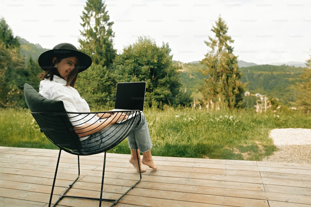 Hipster girl with laptop sitting on wooden porch with beautiful view on woods and mountains. Stylish Young woman in hat using laptop, shopping or working online outside. Freelance and freelancer