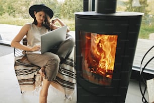 Stylish hipster girl sitting with laptop on cozy chair near fireplace with fire in modern living room with big windows. Working home online. Freelance and freelancer concept. Leisure time