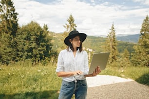 Hipster girl using laptop on background of beautiful view on woods and mountains. Stylish young woman in hat blogging, shopping or working online outside. Travel and Freelance.