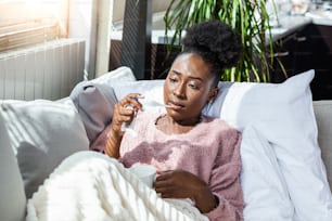 Sickness, seasonal virus problem concept. Woman being sick having flu lying on sofa looking at temperature on thermometer. Sick woman lying in bed with high fever. Cold flu and migraine.