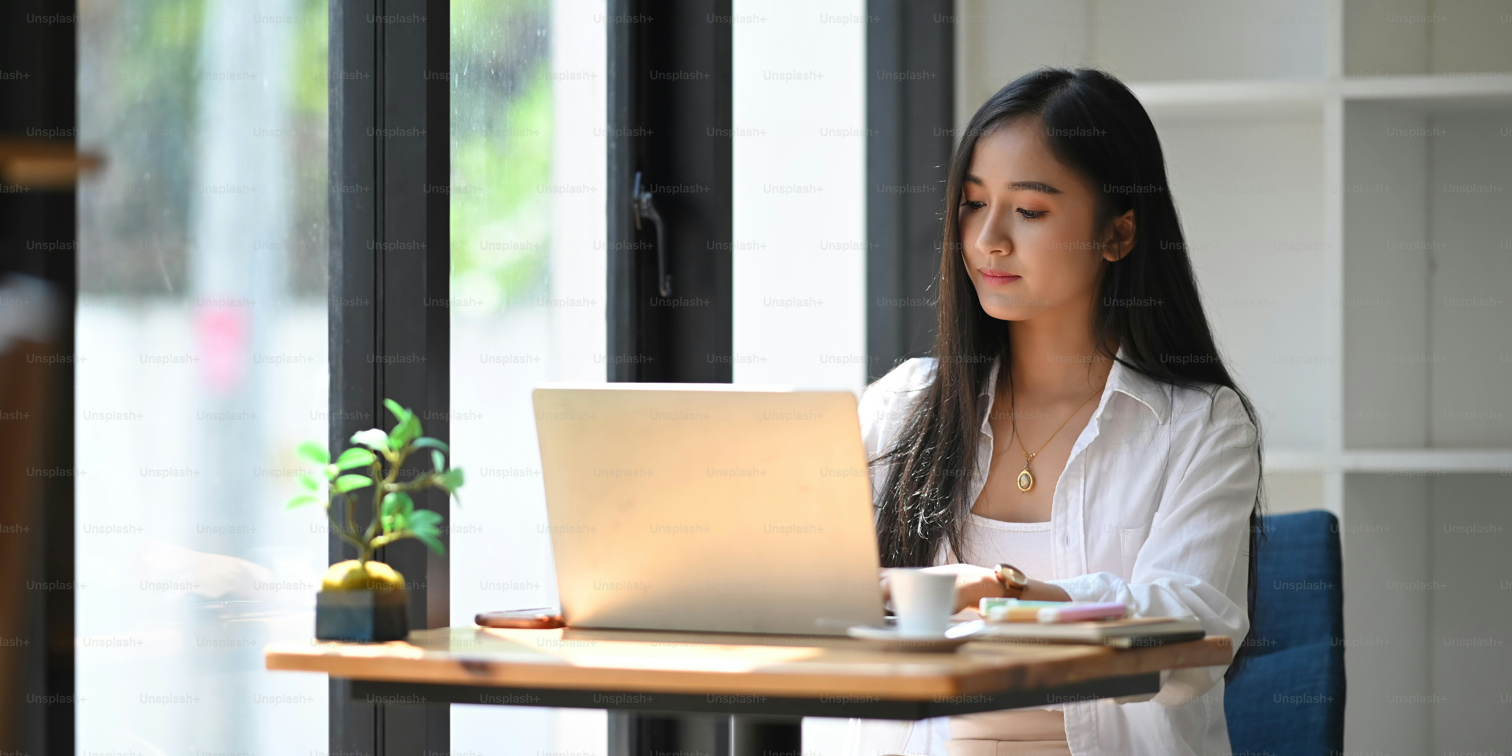 Creative woman using/typing on computer laptop that putting on wooden working desk with coffee cup, notebook, marker pens and potted plant. Work at home concept.