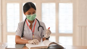 Photo of young woman working as doctor wearing a stethoscope and sitting at the wooden table with book and computer tablet over orderly doctor's office as background.
