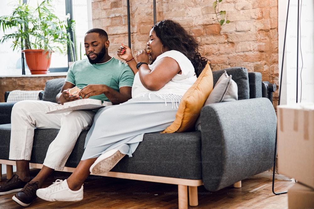 Couple afro-américain assis sur un canapé à la maison et mangeant de la pizza