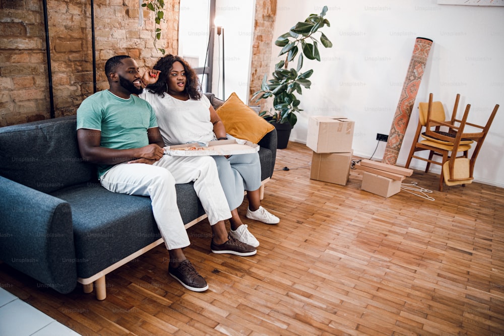 Jeune couple afro-américain assis sur le canapé avec une pizza après avoir emménagé dans un nouvel appartement