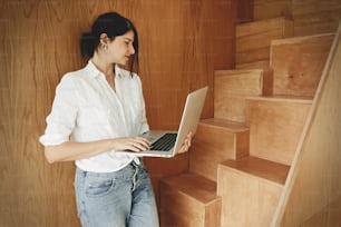 Happy stylish girl using laptop, standing at wooden stairs in modern room. Young casual woman shopping online or working online from home and smiling. Stay home. Freelance