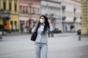 若くてエレガントなビジネスウーマンが誰もいない街の通りを歩き、危険なインフルエンザやウイルスから身を守るために保護マスクを着用しています。コロナウイルスまたはCovid-19のコンセプト。