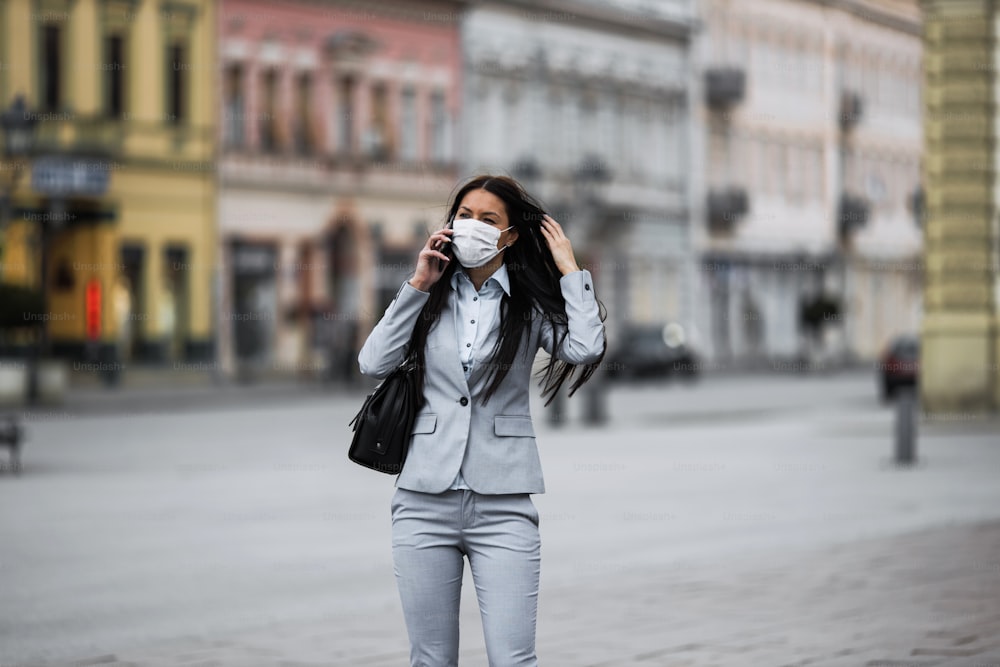 Giovane ed elegante donna d'affari che cammina sulla strada vuota della città e indossa una maschera protettiva per proteggersi da influenze o virus pericolosi. Concetto di virus corona o Covid-19.