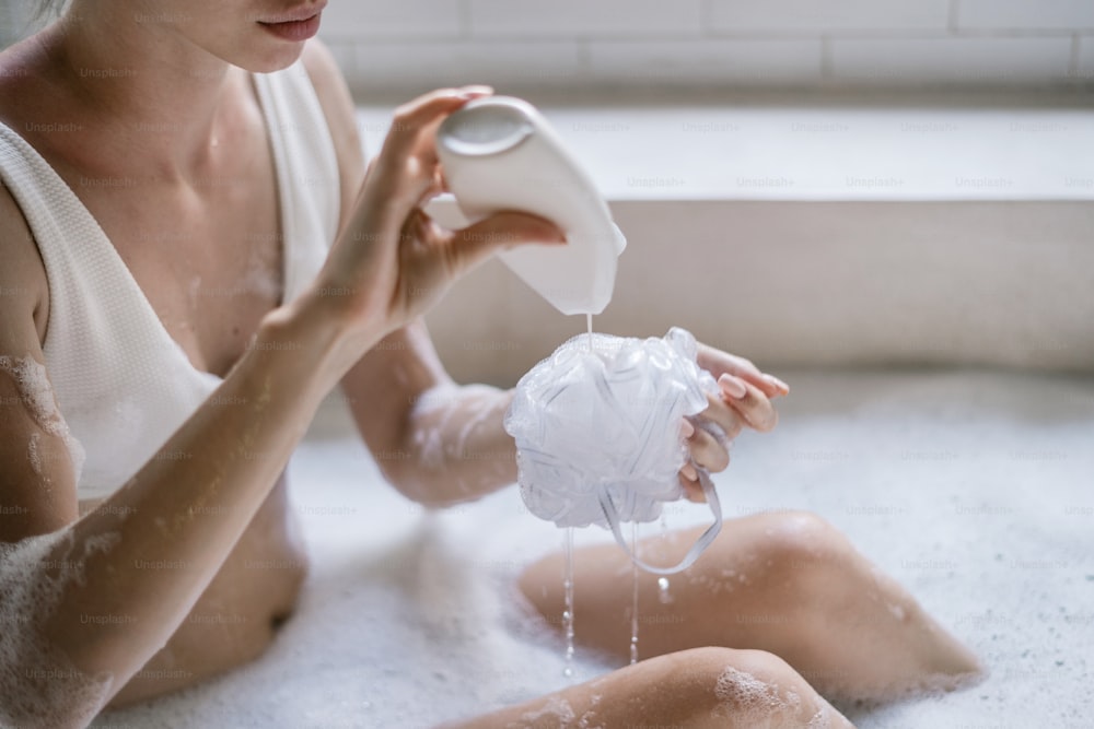 Conceito de cuidados com o corpo. Vista recortada da mulher adulta jovem derramando gel de banho na esponja, tomando banho, deitada em água de espuma de sabão em casa