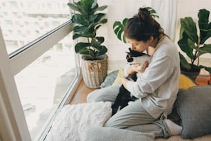 Chica hipster abrazando a un lindo gato, sentados juntos en casa durante la cuarentena por coronavirus. Quédate en casa, mantente a salvo. Aislamiento en casa para prevenir la epidemia del virus. Mujer joven con gato en habitación moderna