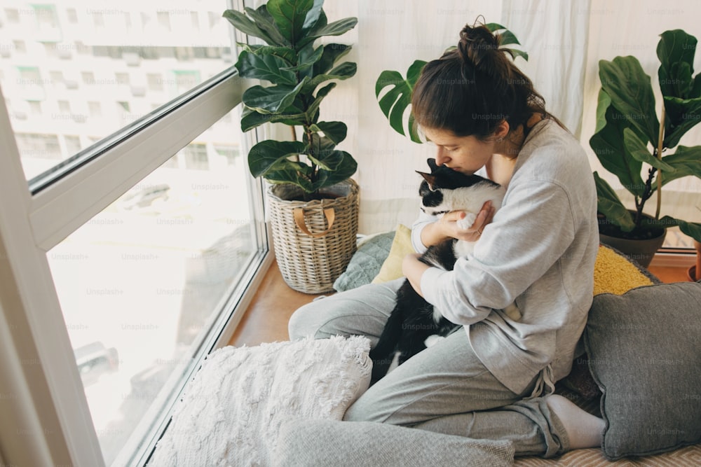 Hipster girl hugging cute cat, sitting together at home during coronavirus quarantine. Stay home stay safe. Isolation at home to prevent virus epidemic. Young woman with cat in modern room