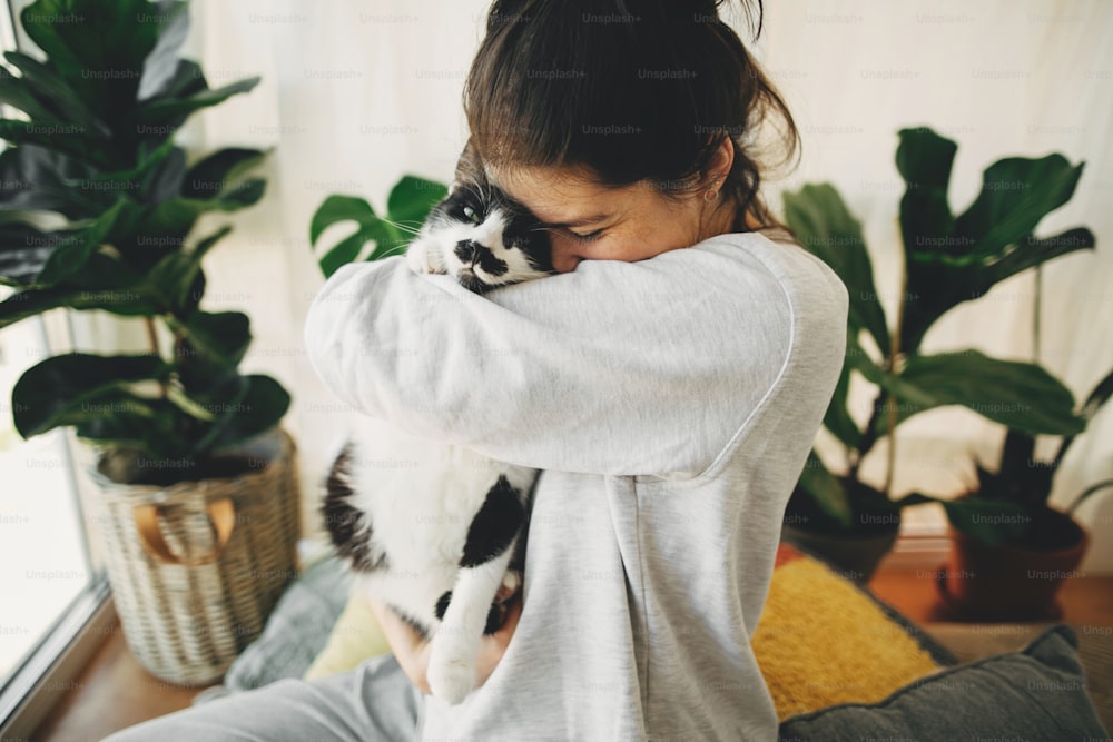 かわいい猫を抱きしめ、コロナウイルスの検疫中に家で一緒に座っている幸せなヒップスターの女の子。家にいて、安全を確保してください。ウイルスの流行を防ぐための自宅での隔離。モダンな部屋に猫を連れた若い女性