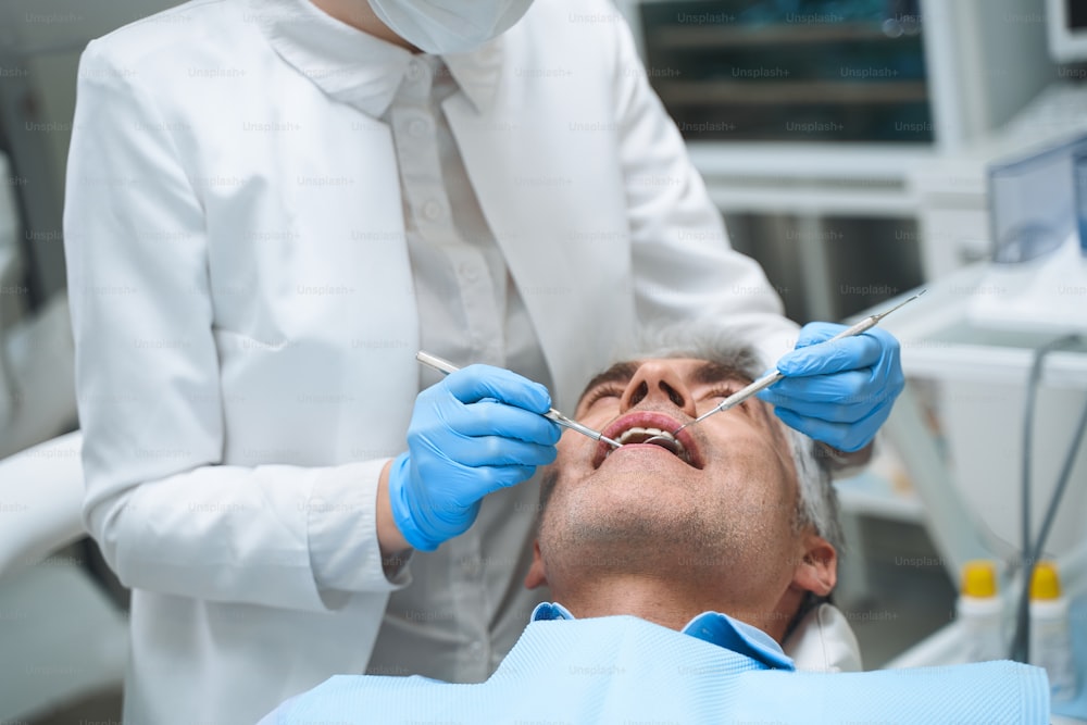 Mann liegt im Zahnarztstuhl, während der Spezialist ihn mit Spiegel und Entdecker untersucht