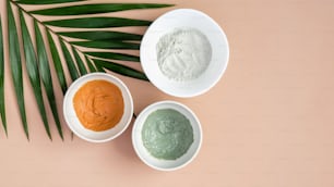 SPA cosmetic facial masks in bowls and tropical leaf on peach background. Face skin care and beauty treatment concept. Flat lay, top view