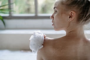 スキンケアのコンセプト。穏やかな若い成人女性が入浴し、スポンジを手に持ち、肩を洗い、バスルームで朝を過ごす後ろ姿