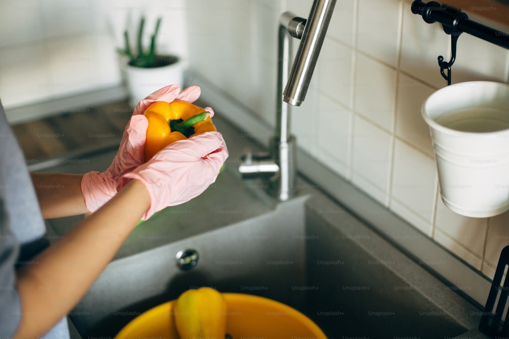 Mãos em luvas segurando pimenta, lavando legumes durante a epidemia de vírus. Mulher nas mãos cor-de-rosa lavando legumes frescos, preparando para cozinhar a refeição na cozinha moderna