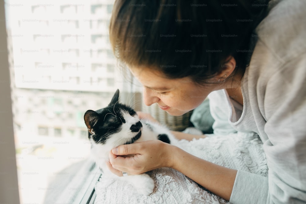 かわいい猫を抱きしめ、コロナウイルスの検疫中に家で一緒に座っているヒップスターの女の子。家にいて、安全を確保してください。ウイルスの流行を防ぐための自宅での隔離。モダンな部屋に猫を連れた若い女性