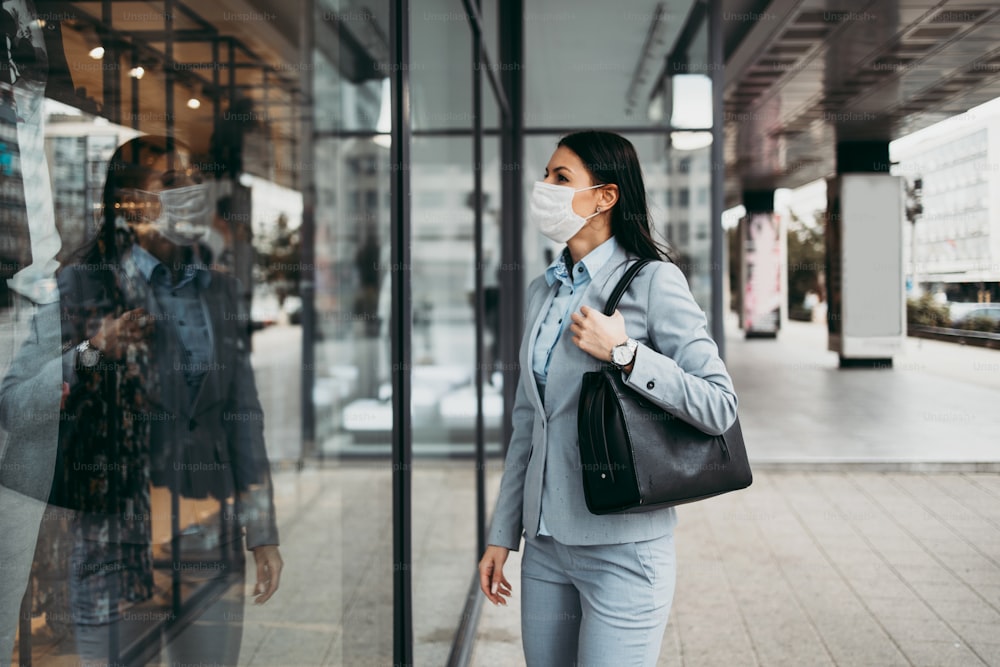 Junge und elegante Geschäftsfrau, die auf die Ladenfront in der leeren Stadtstraße schaut und eine Schutzmaske trägt, um sich vor gefährlichen Grippen oder Viren zu schützen. Coronavirus oder Covid-19-Konzept.