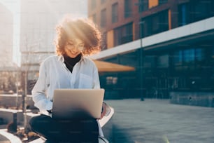 Smiling curly haired caucasian woman working outside on her computer while sitting on stone stairs against the sunshine