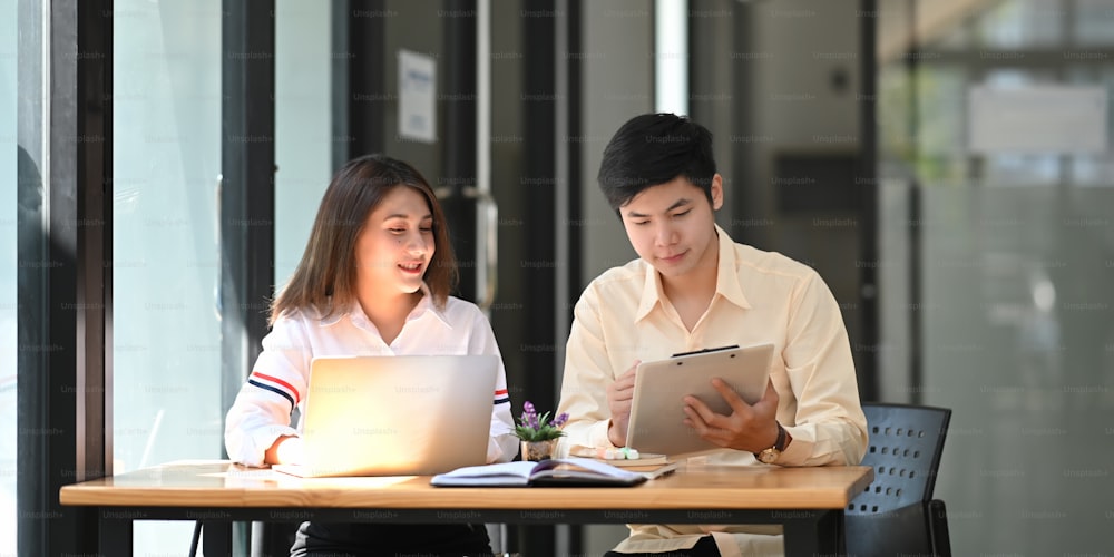 Photo of young business developer team discussing/meeting/working with computer laptop and tablet while sitting together at wooden working desk over orderly living room as background.