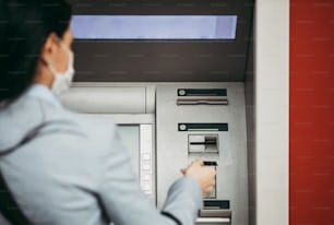 Elegant business woman with protective mask standing on city street and using ATM machine to withdraw cash. Corona or Covid-19 virus pandemic concept.