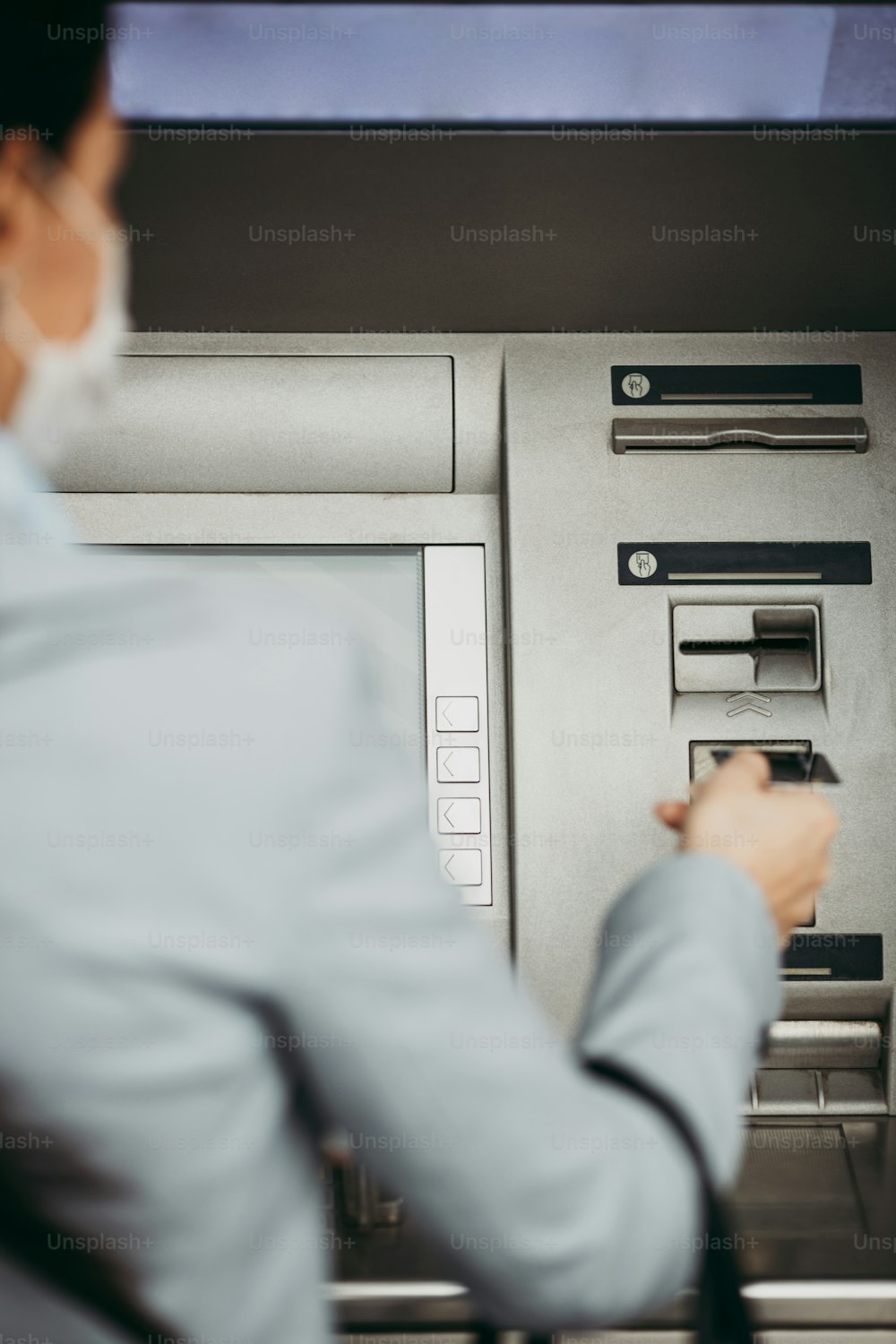 Elegant business woman with protective mask standing on city street and using ATM machine to withdraw cash. Corona or Covid-19 virus pandemic concept.