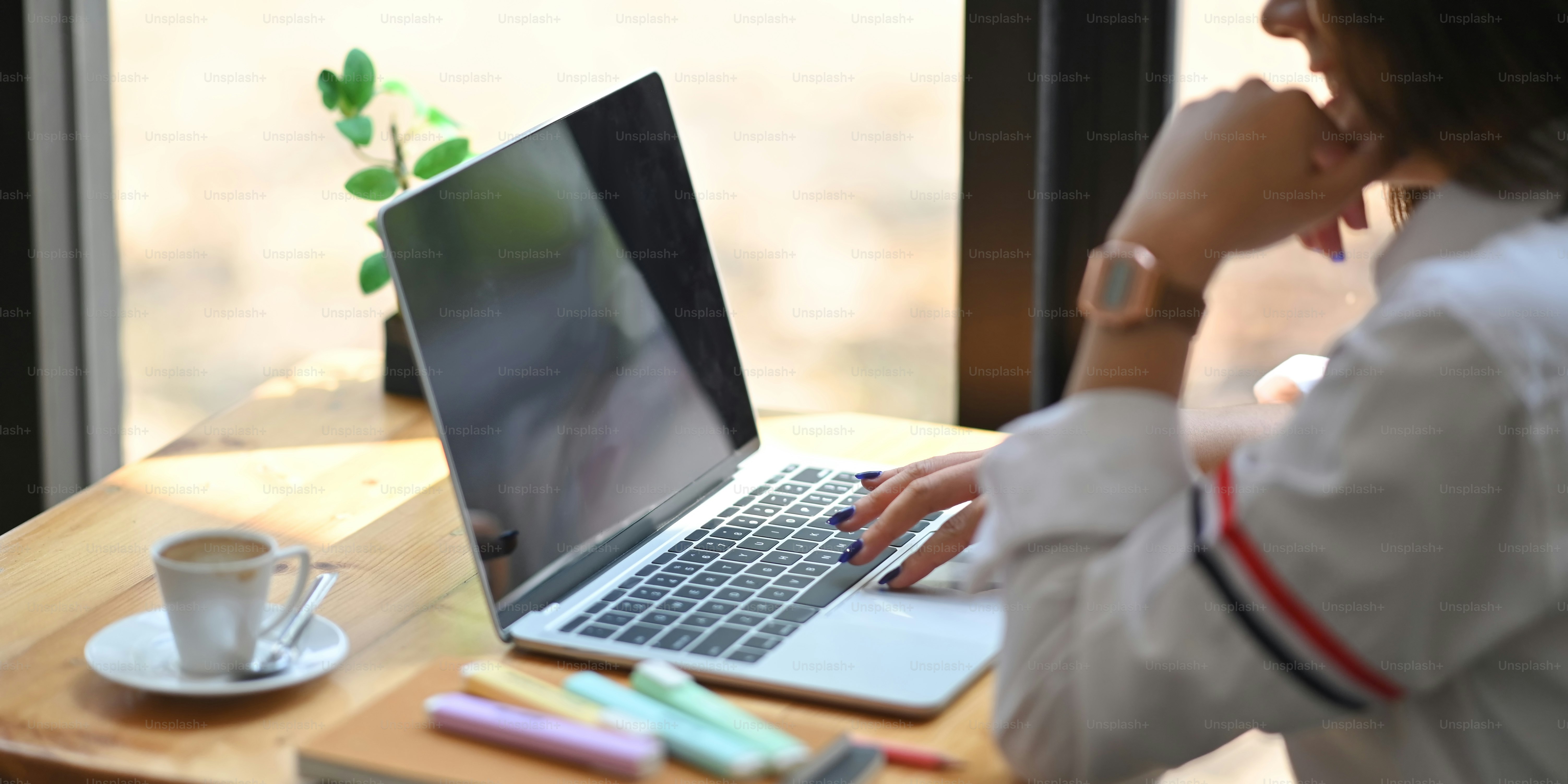 Cropped shot woman using/typing on computer laptop that putting on wooden working desk with coffee cup, notebook, marker pens and potted plant. Work at home concept.