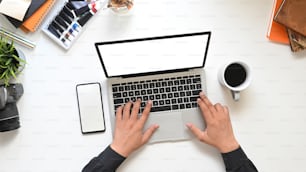 上面図 コーヒーカップ、空の画面、スマートフォン、アクセサリーに囲まれた白い作業台に置く白い空白の画面を備えたコンピューターのラップトップで入力するクリエイティブな男性の手。